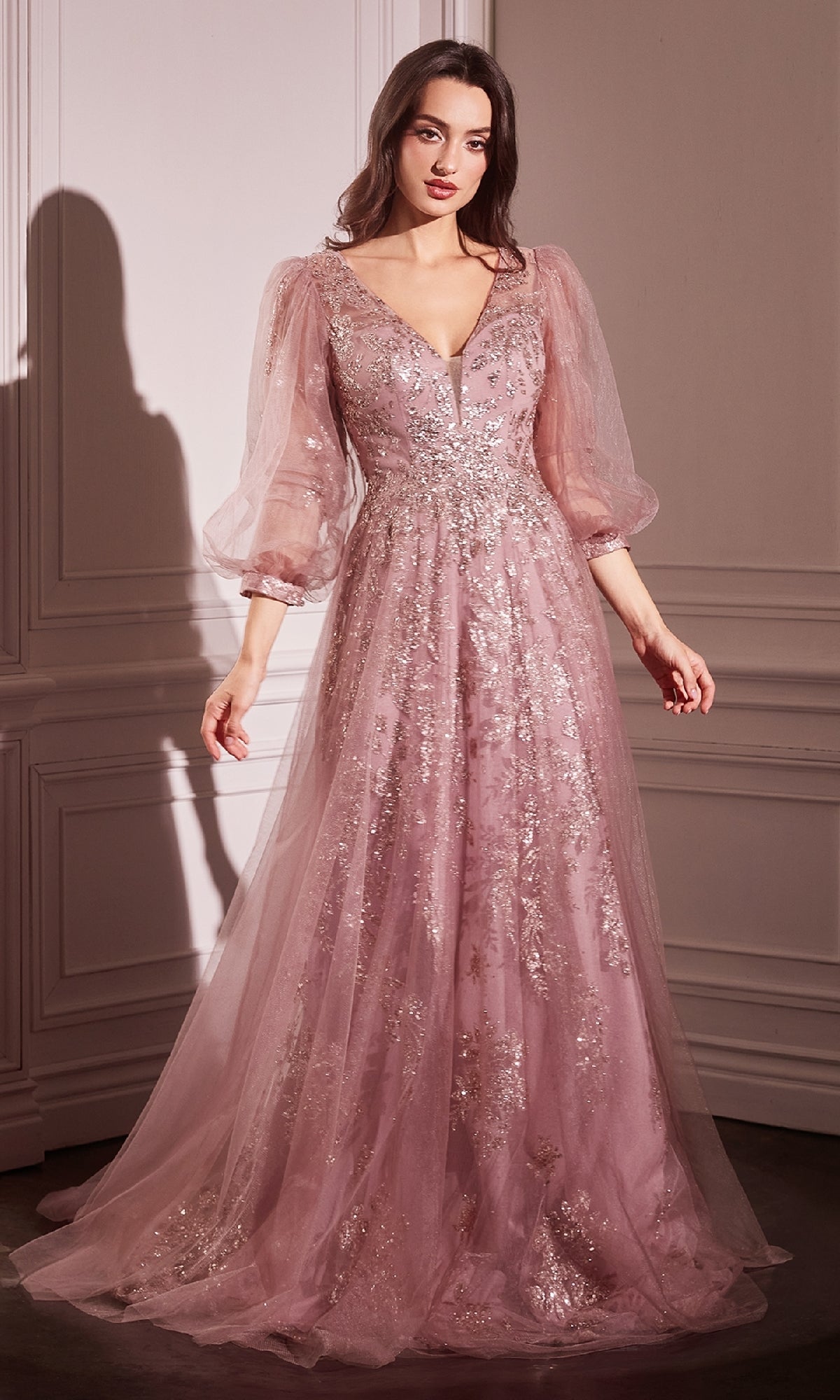 A woman wears the Ladivine CD0102 Sheer-Sleeve Long Prom Dress, adorned with silver sequins and sheer puff sleeves, standing in a softly lit, elegant room.