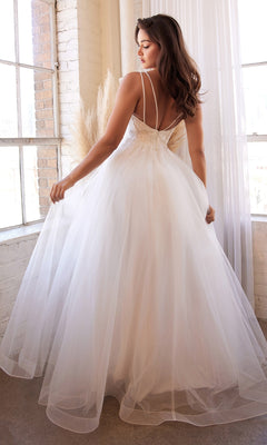 A woman stands indoors by large windows in the Off-White Long A-Line Wedding Dress CD0154W, featuring thin straps, an open back, and a flowing tulle design for an elegant, timeless bridal look.