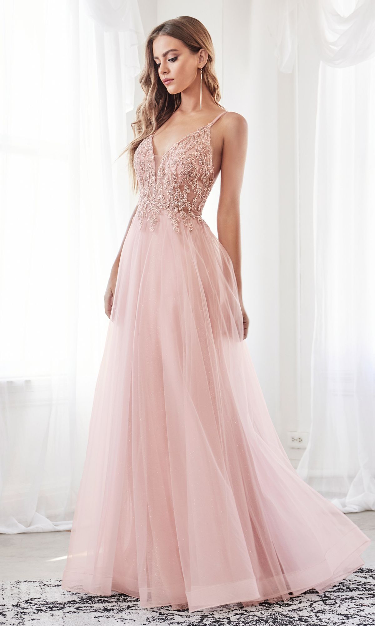 A woman stands indoors by a window wearing the Sheer-Corset Long Prom Ball Gown CD0154, a floor-length blush pink dress featuring a lace bodice, beaded accents, and a flowing tulle skirt.