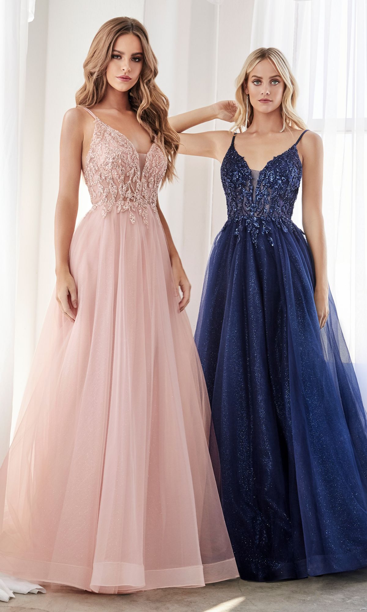 Two women stand indoors in floor-length formal dresses; one wears a light pink gown with lace and beads, while the other dons the Sheer-Corset Long Prom Ball Gown CD0154—a dark blue sequin dress with a tulle skirt.