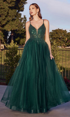 A woman stands outdoors in the Sheer-Corset Long Prom Ball Gown CD0154, a floor-length deep green dress with beaded accents, spaghetti straps, a fitted bodice, and a full tulle skirt, framed by lush trees and bushes.