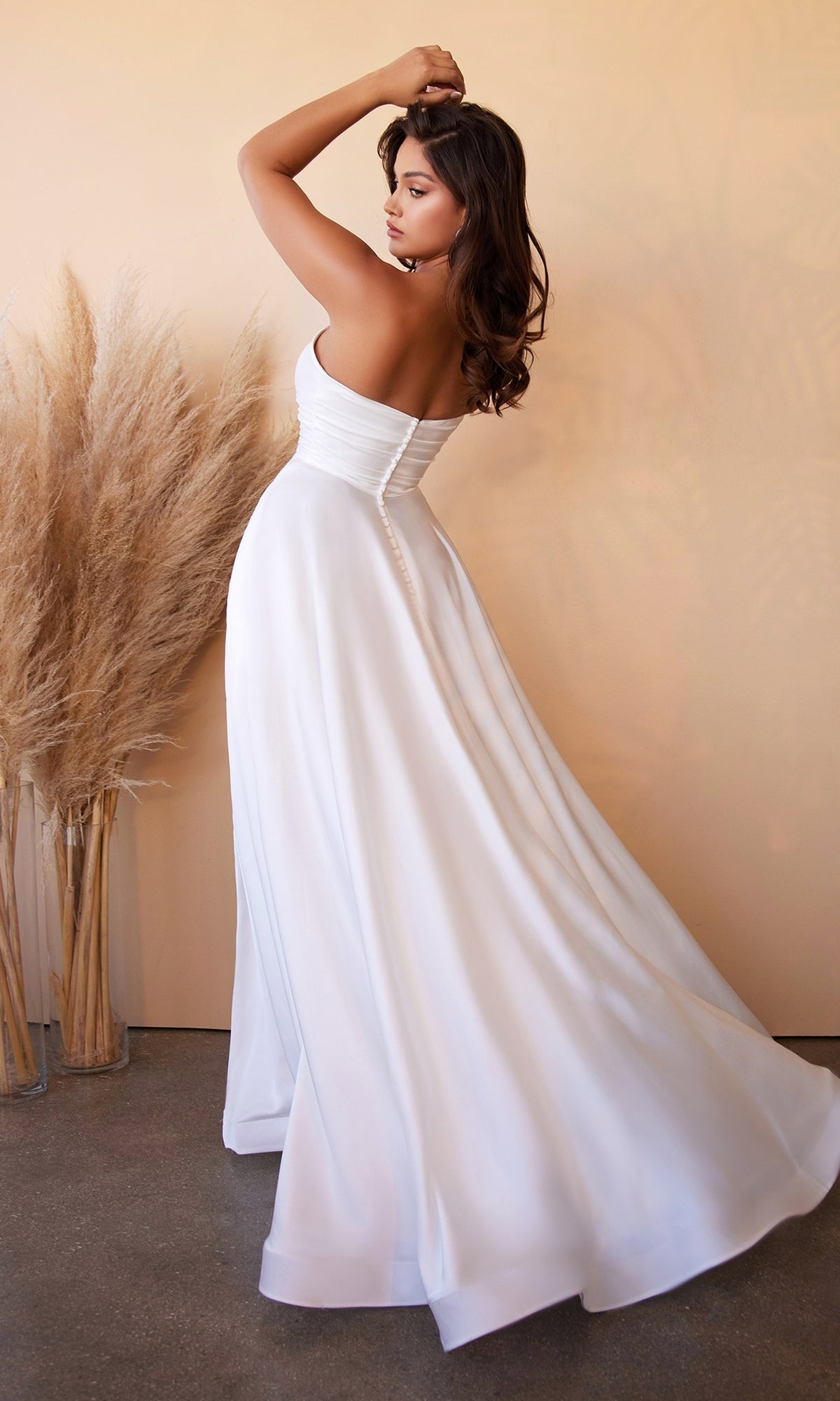 A woman wearing the Strapless Long White Satin Formal Dress CD0166W poses with one arm raised and her back to the camera, standing in front of a beige wall adorned with pampas grass.