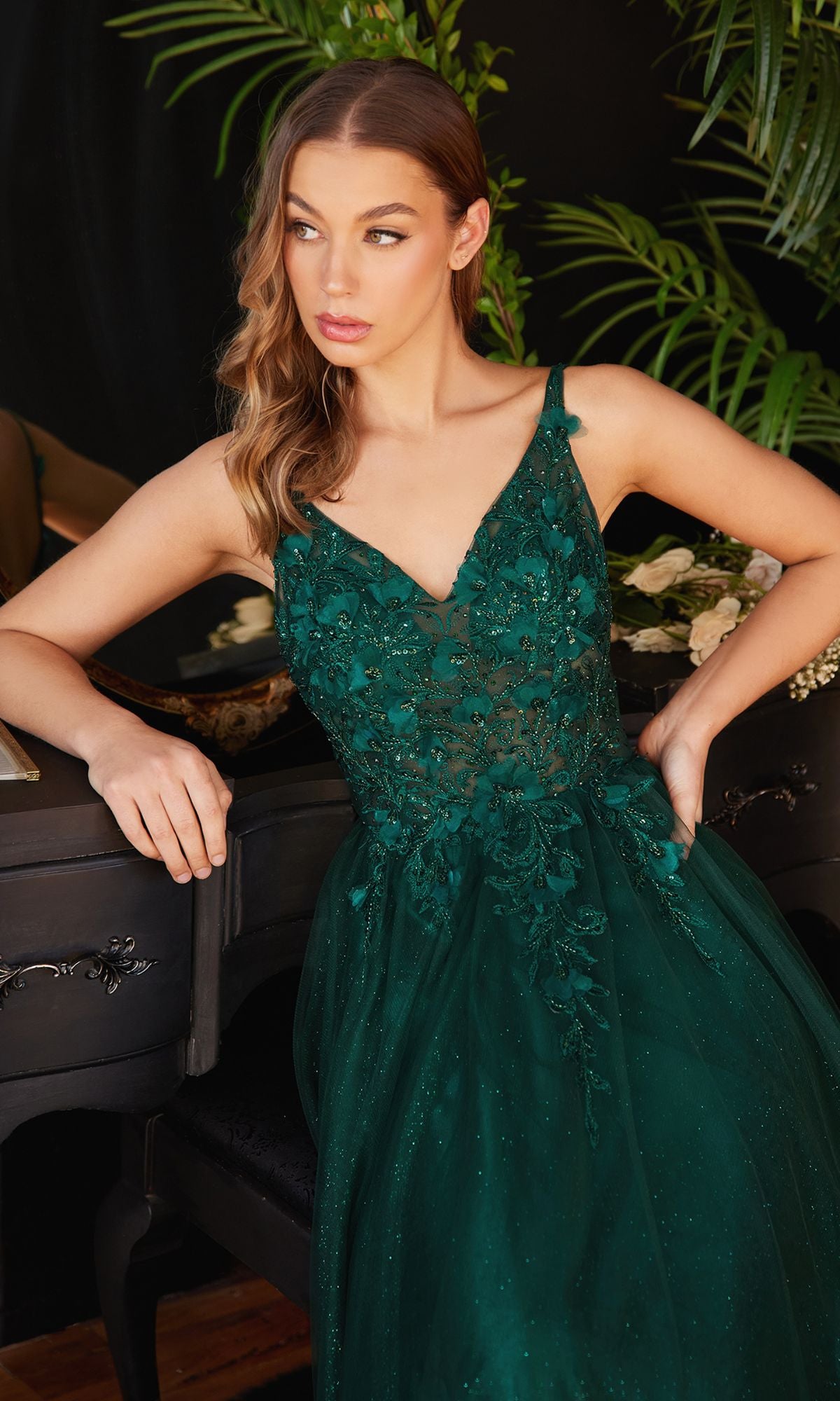 A woman in the Glitter Tulle Illusion Prom Dress: Ladivine CD0181, dark green and embroidered, sits by a black piano adorned with flowers, surrounded by lush green plants.