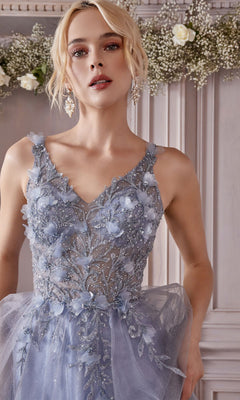A woman in the Glitter Tulle Illusion Prom Dress: Ladivine CD0181, featuring light blue embroidery and sheer details, stands before a light ornate wall with white flowers and greenery in the background.