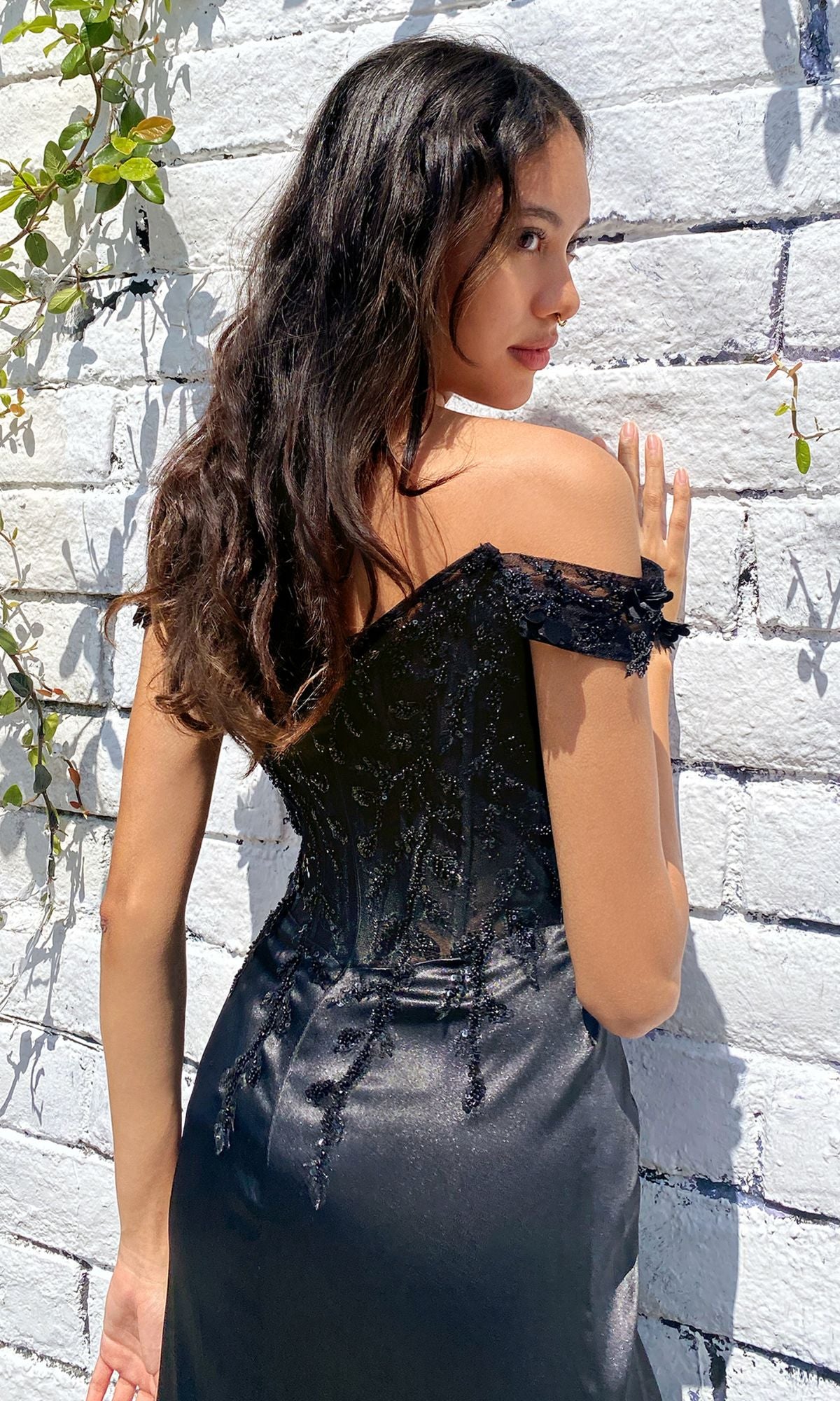 A woman with long brown hair models the Off-the-Shoulder Long Sweetheart Prom Dress CD0186, featuring glittery embroidery, while standing by a white brick wall decorated with green vines.