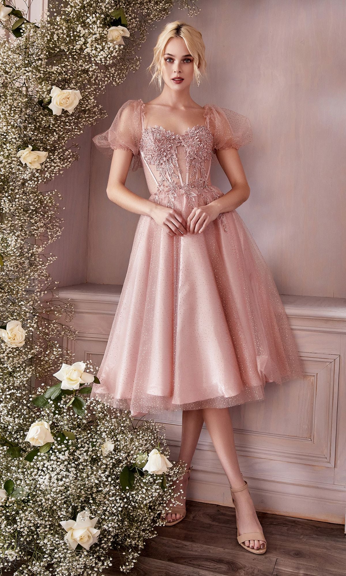 Wearing the blush pink, sparkly Puff-Sleeve Tea-Length Prom Dress: CD0187, a woman stands beside a floral arrangement of white roses and baby's breath against a pale wall.