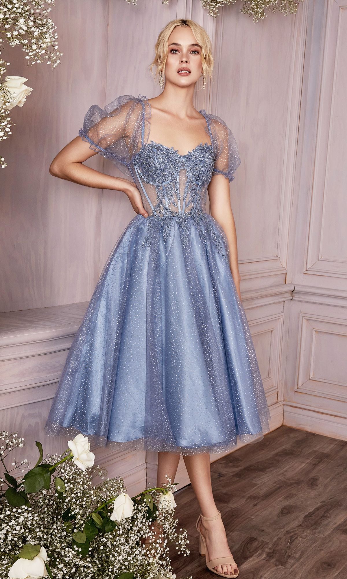 A woman poses indoors in the Puff-Sleeve Tea-Length Prom Dress: CD0187, a light blue dress with sheer tulle, puff sleeves, and floral details, surrounded by white flowers and wooden panel walls.
