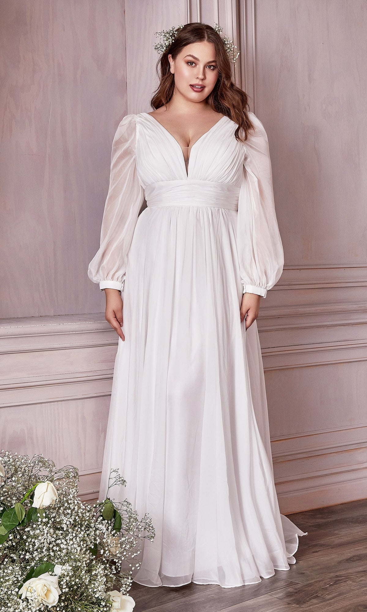 A woman stands indoors in a Long White Chiffon Formal Dress with Long Sleeves, under $200, paired with floral hair accessories beside a bouquet of white flowers.