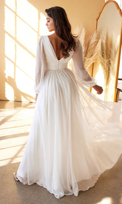 A woman stands indoors facing away, sunlight streaming through a window and reflecting in a large mirror. She wears the Long White Chiffon Formal Dress with Long Sleeves—a flowing, elegant option perfect as an affordable wedding dress under $200.