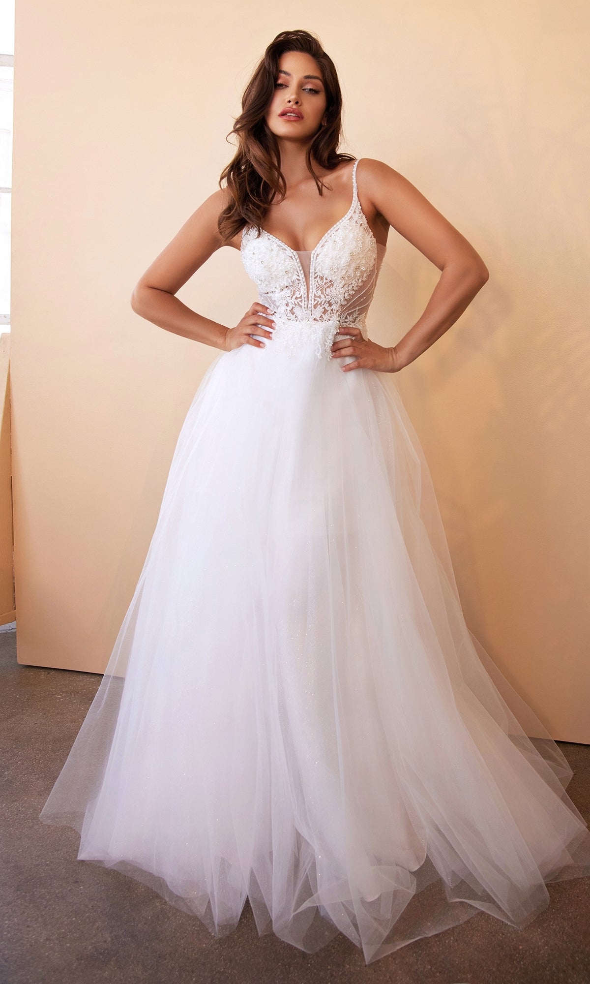 A woman stands against a beige wall wearing the Sheer-Bodice Long White Ball Gown CD0195W—a sleeveless bridal dress with tulle skirt, deep neckline, and fitted bodice.