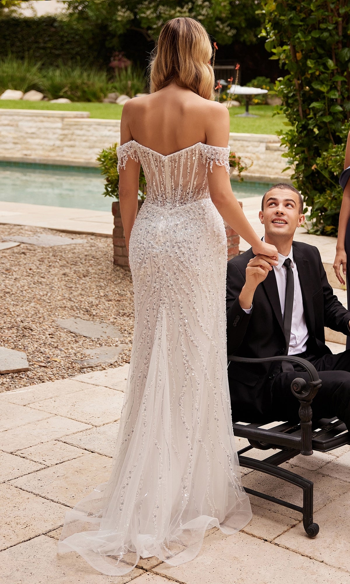A woman wears the Long White Off-Shoulder Sequin Prom Dress CD0203W, standing beside a man in a black suit seated outdoors near a pool.