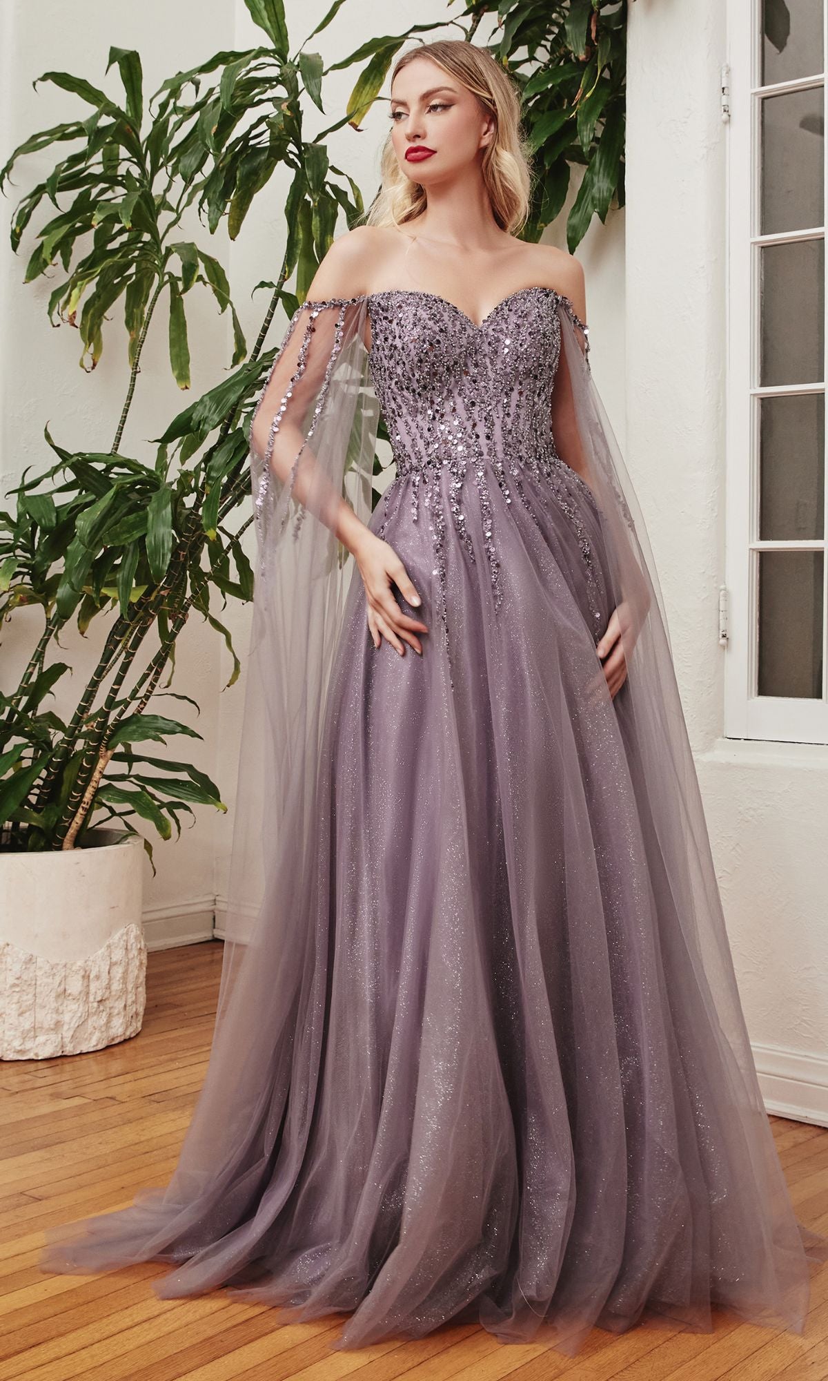 A woman stands indoors wearing the Cape-Sleeve Long Beaded Prom Dress CD0204, a floor-length lavender A-line gown with beaded embellishments and sheer cape sleeves.