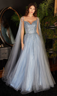 A woman stands indoors by a mirror and greenery, wearing the Cape-Sleeve Long Beaded Prom Dress CD0204—a light blue, floor-length gown with sheer tulle cape sleeves.