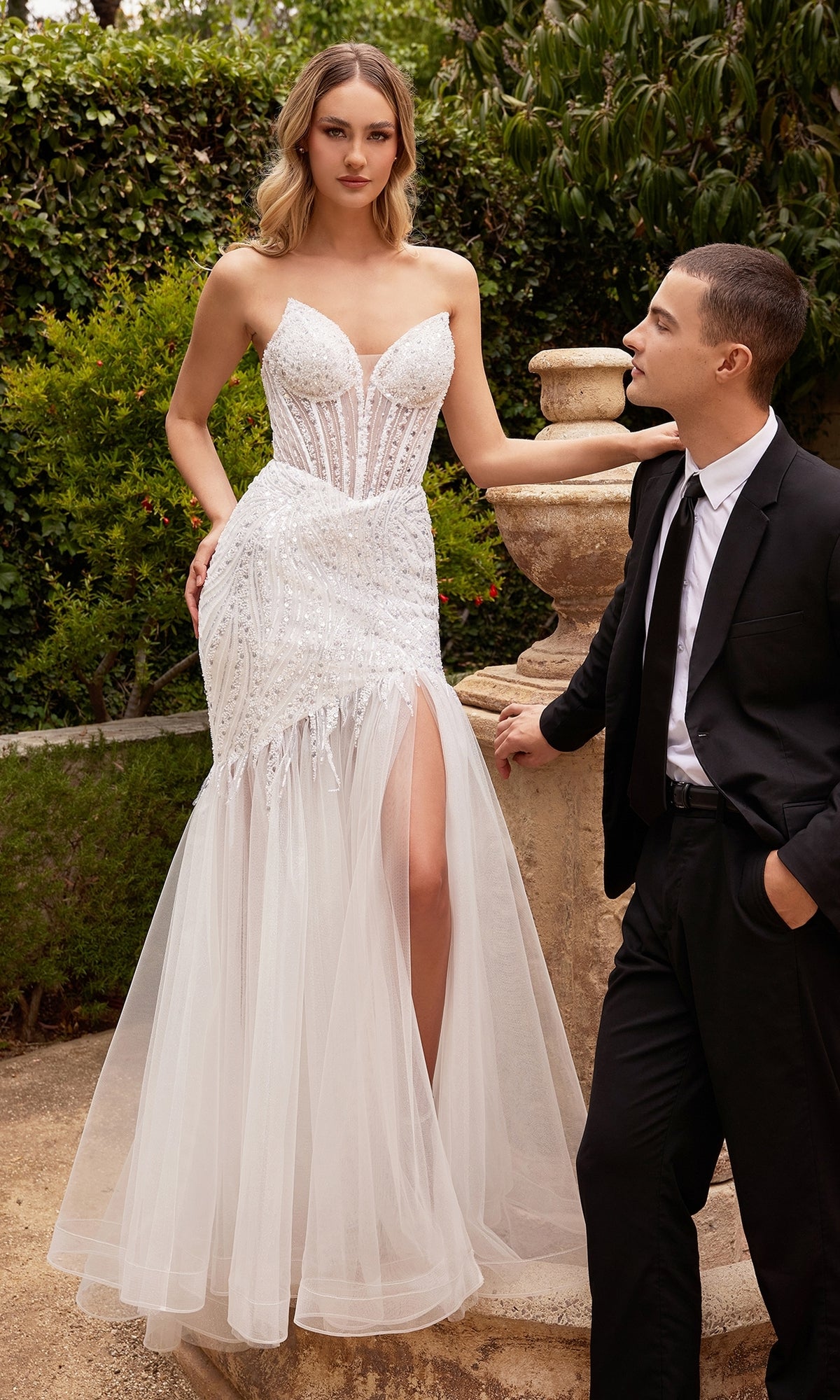 A woman in the Strapless Long White Mermaid Wedding Gown CD0215W stands next to a man in a black suit outdoors, her elegant white dress shimmering with embellishments and featuring a tulle skirt and thigh-high slit.