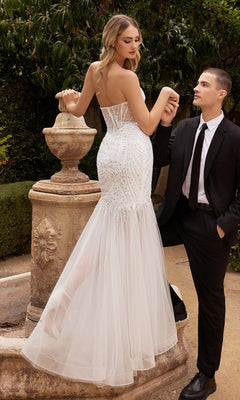 A woman wearing the Strapless Long White Mermaid Wedding Gown CD0215W stands by a stone fountain as a man in a black suit and tie holds her hand and looks at her.