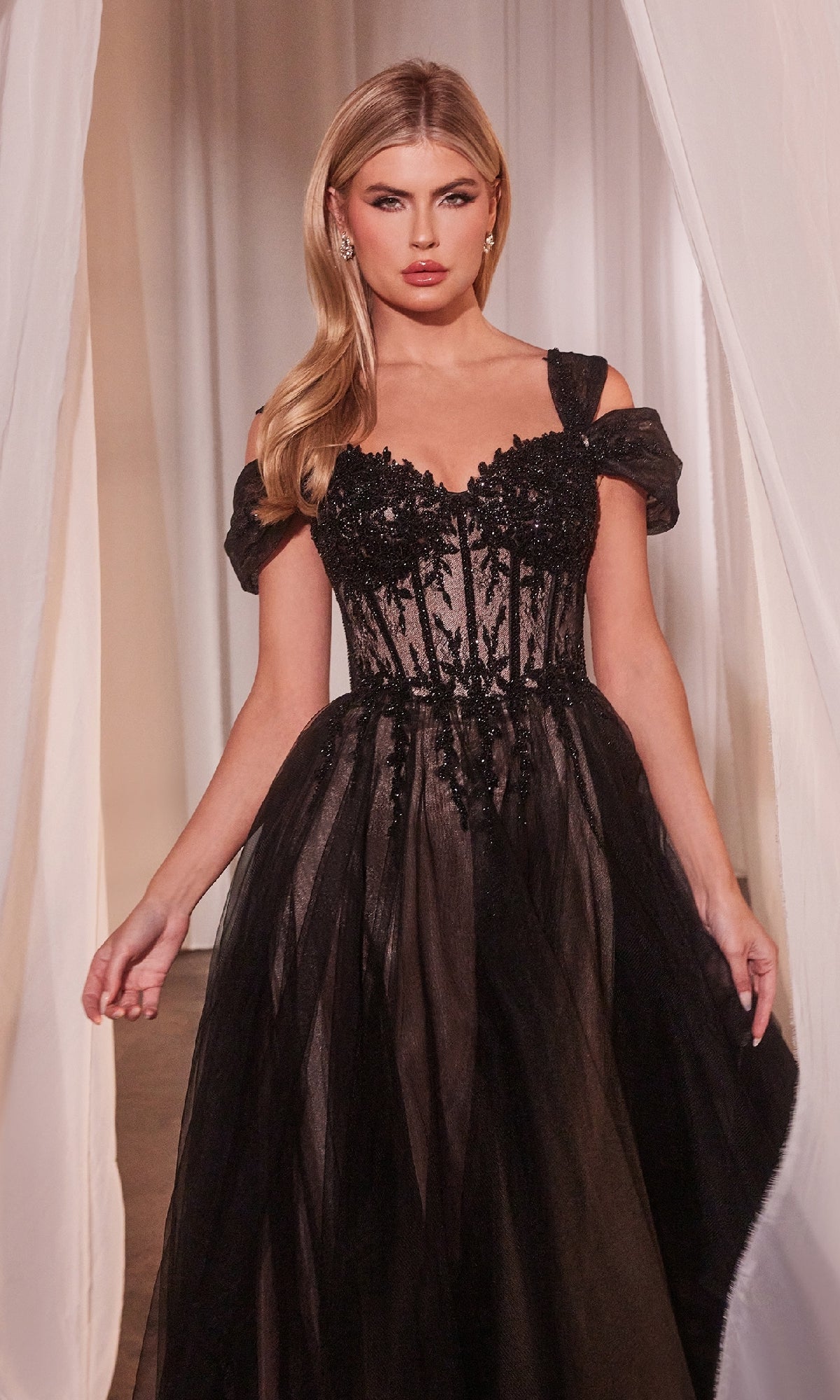A woman in the Lace-Up Long Black Prom Dress: Ladivine CD0237 with a sheer bodice and sequin embellishments stands between white curtains, highlighting the elegance of this formal evening gown.