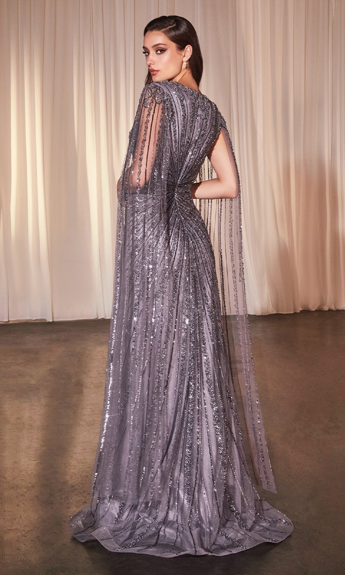 A woman stands before light curtains, looking over her shoulder in the Cape-Sleeve Beaded Formal Dress: Ladivine CD0239, a long gown featuring draped sheer cape sleeves and silver sequins.