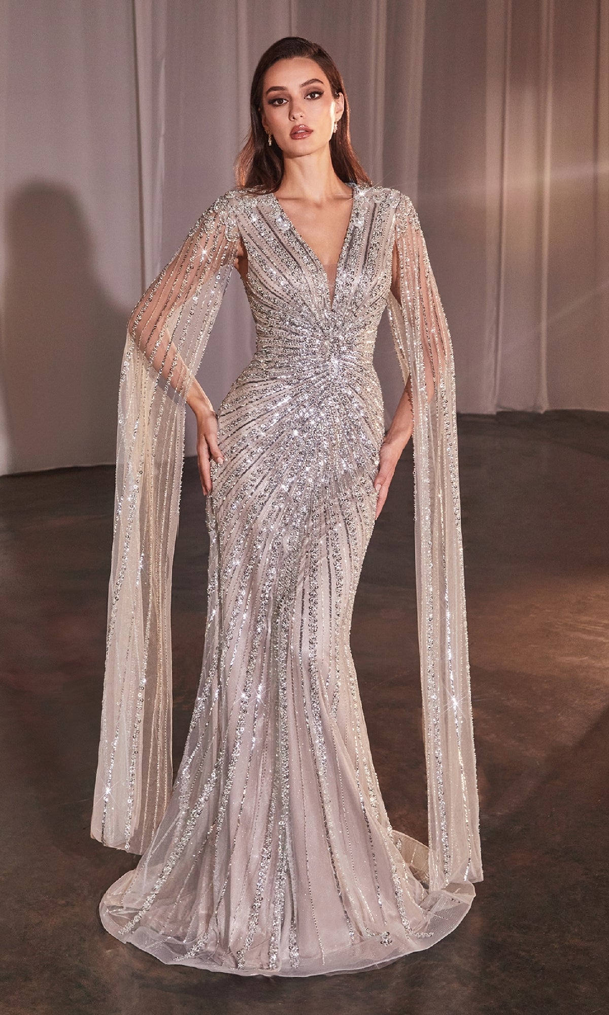 A woman models the Ladivine CD0239 Cape-Sleeve Beaded Formal Dress—a floor-length silver sequin gown with a fitted bodice, deep V-neckline, and sheer cape sleeves—against a simple indoor backdrop.