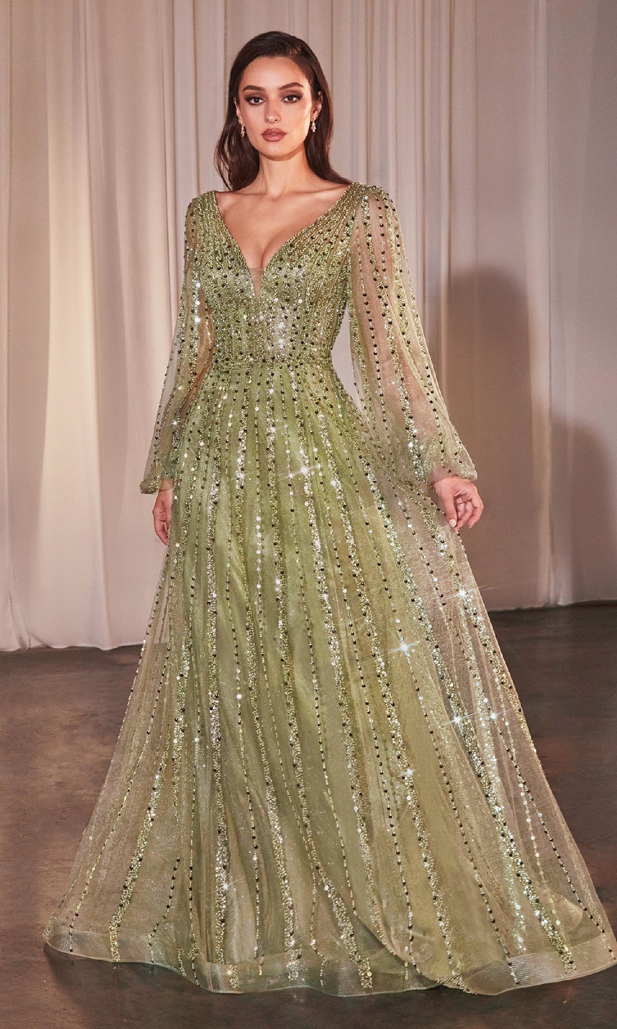 A woman stands indoors on a hardwood floor with light curtains behind her, wearing the Ladivine CD0240—a long-sleeve, floor-length olive green formal dress adorned with sequins.