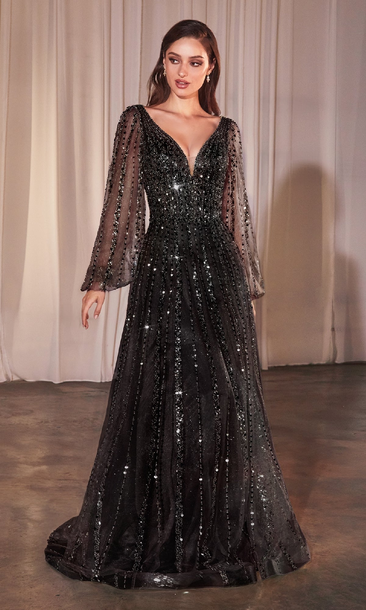 A woman stands indoors in the Ladivine CD0240, a floor-length black formal dress with sheer long sleeves and sparkling sequin beading, set against white curtains.