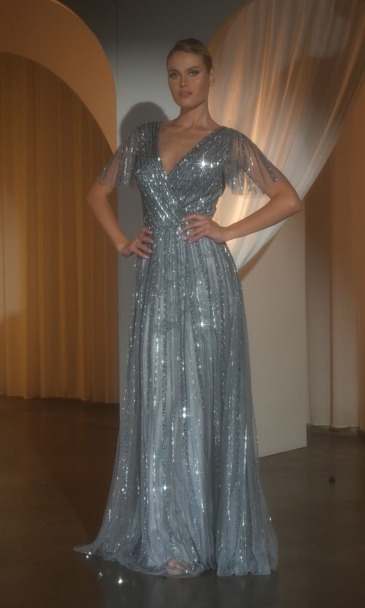 A woman stands indoors wearing the Ladivine CD0241 Flutter-Sleeve Long Prom Dress, a floor-length shimmering silver gown with short, sheer sleeves and elegant sequin detailing.