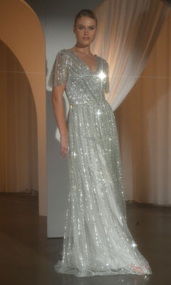 A woman stands indoors wearing the Ladivine CD0241 Flutter-Sleeve Long Prom Dress—a floor-length, silver sequin gown with sheer overlay—while light reflects off its surface and curtains appear in the background.