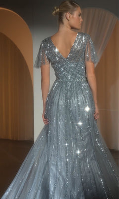 A blonde-haired woman in a bun wears the Ladivine CD0241 Flutter-Sleeve Long Prom Dress, a sparkly silver gown with a V-back and sheer embellished flutter sleeves, standing indoors.