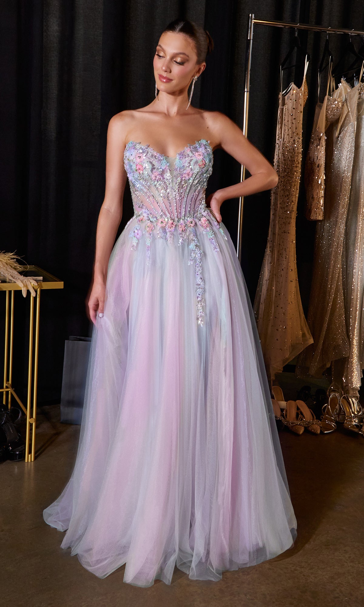 A woman stands in a backstage area, surrounded by evening dresses and shoes, wearing the Beaded-Corset Prom Ball Gown: Ladivine CD2418—a strapless long tulle ball gown in delicate pastel shades with floral embellishments.