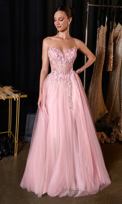 A woman stands indoors near a display of gowns and accessories, wearing the Beaded-Corset Prom Ball Gown: Ladivine CD2418 in light pink, which features a strapless silhouette with a fitted, beaded bodice and flowing tulle skirt. Nearby, a stunning sage green prom dress and an elegant strapless long tulle prom dress are also showcased.