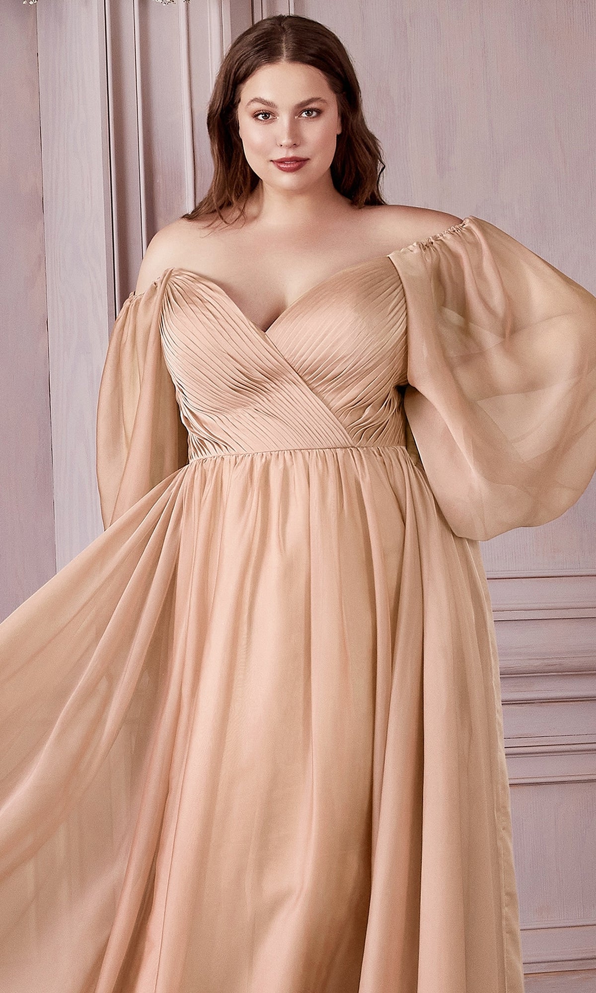 A woman stands before a light wooden wall wearing the Ladivine Plus-Size Off-Shoulder Prom Dress CD243C in blush, featuring a pleated bodice and sheer, billowy long sleeves.