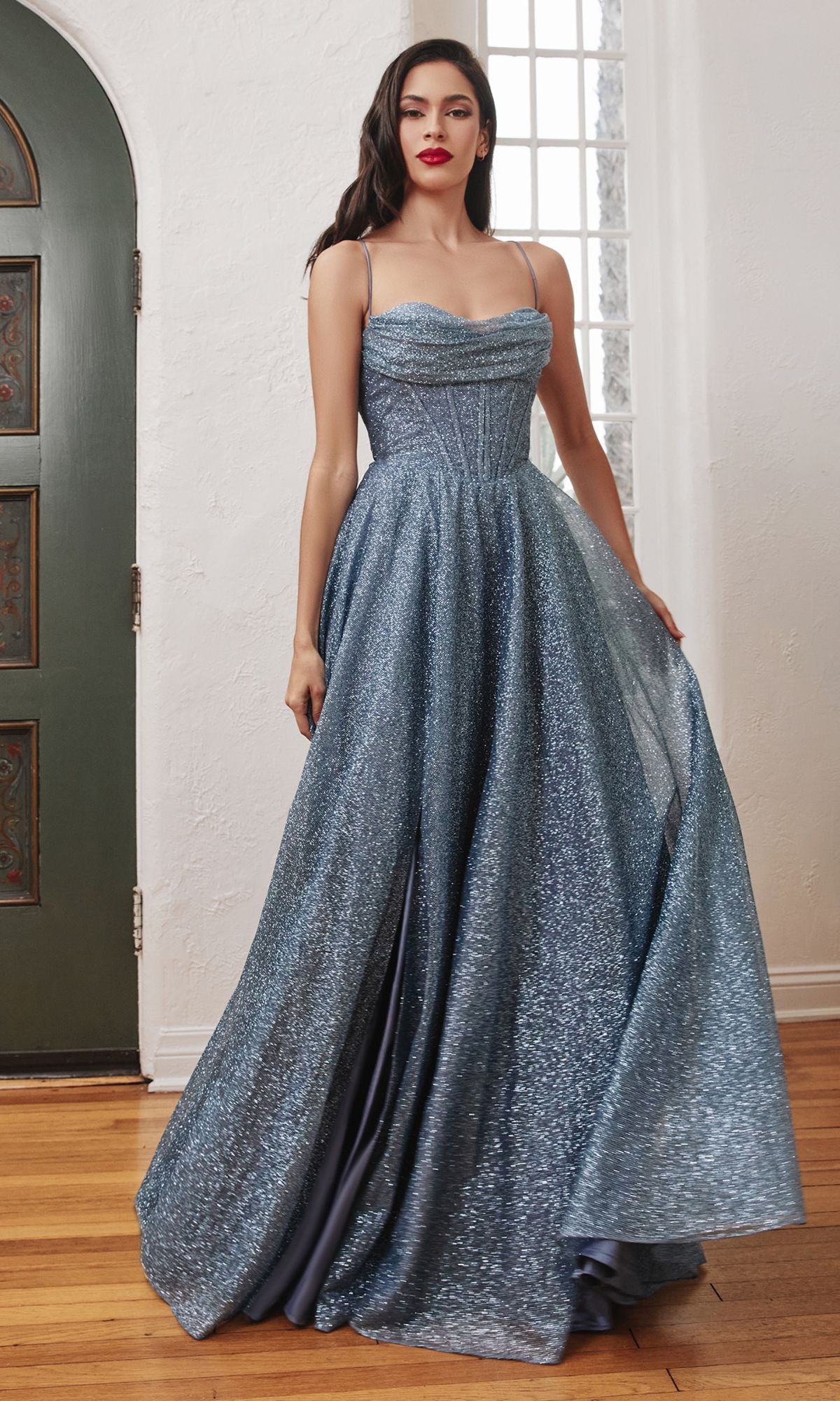 A woman stands indoors near a green door and window, wearing the Draped-Bodice Long Glitter A-Line Prom Dress CD252 in sparkly blue with thin straps and a corset bodice.