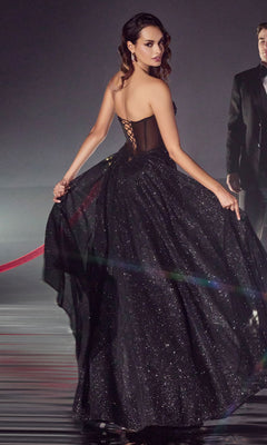 A woman in the Strapless Sheer-Corset Glitter Prom Ball Gown CD275, dazzling and floor-length with a lace-up back, looks over her shoulder while holding the skirt. A man in a tuxedo is visible in the background.