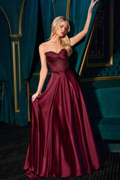 A woman in a burgundy satin Ladivine CD297 strapless long A-line prom dress stands in an elegant room with teal curtains and gold accents.