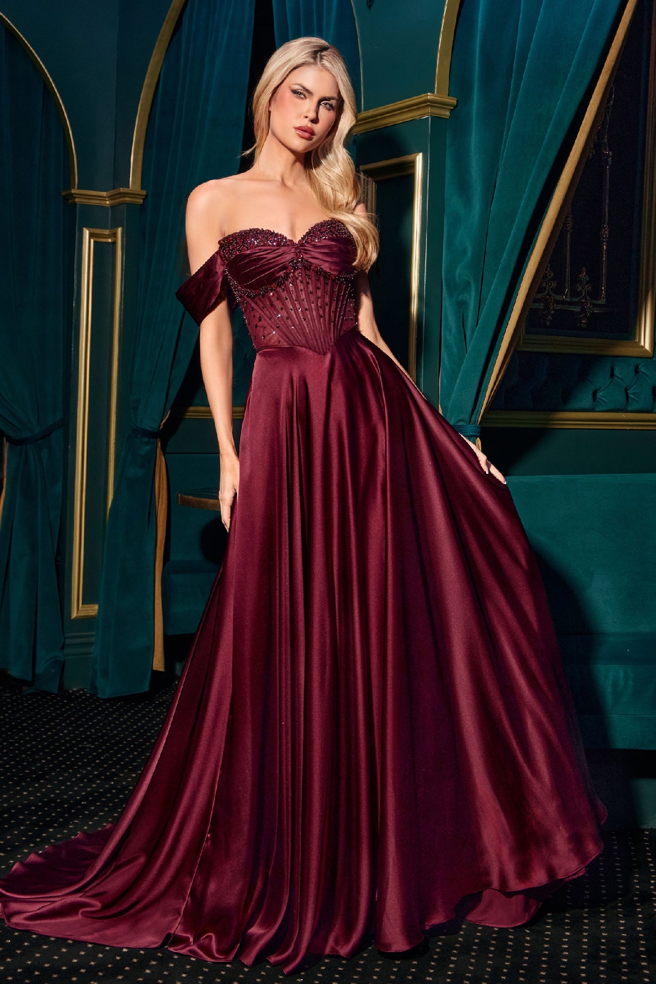 A woman in a Ladivine CD297 Strapless Long A-Line Prom Dress stands in an elegant room with green velvet curtains and gold accents, wearing a burgundy satin off-the-shoulder gown.