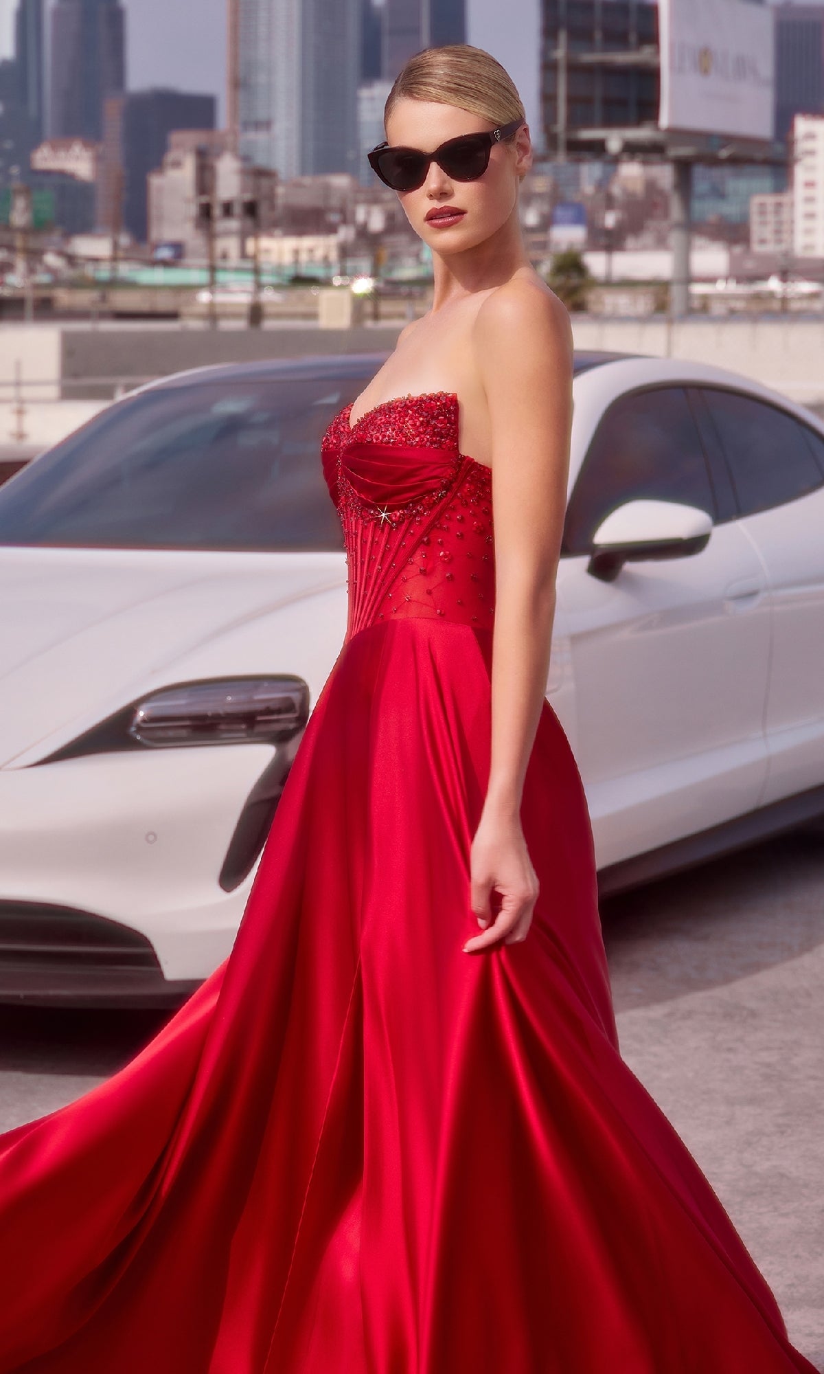 A woman in the Ladivine CD297 Strapless Long A-Line Prom Dress and sunglasses stands confidently before a white car, radiating urban elegance in her chic formal style.
