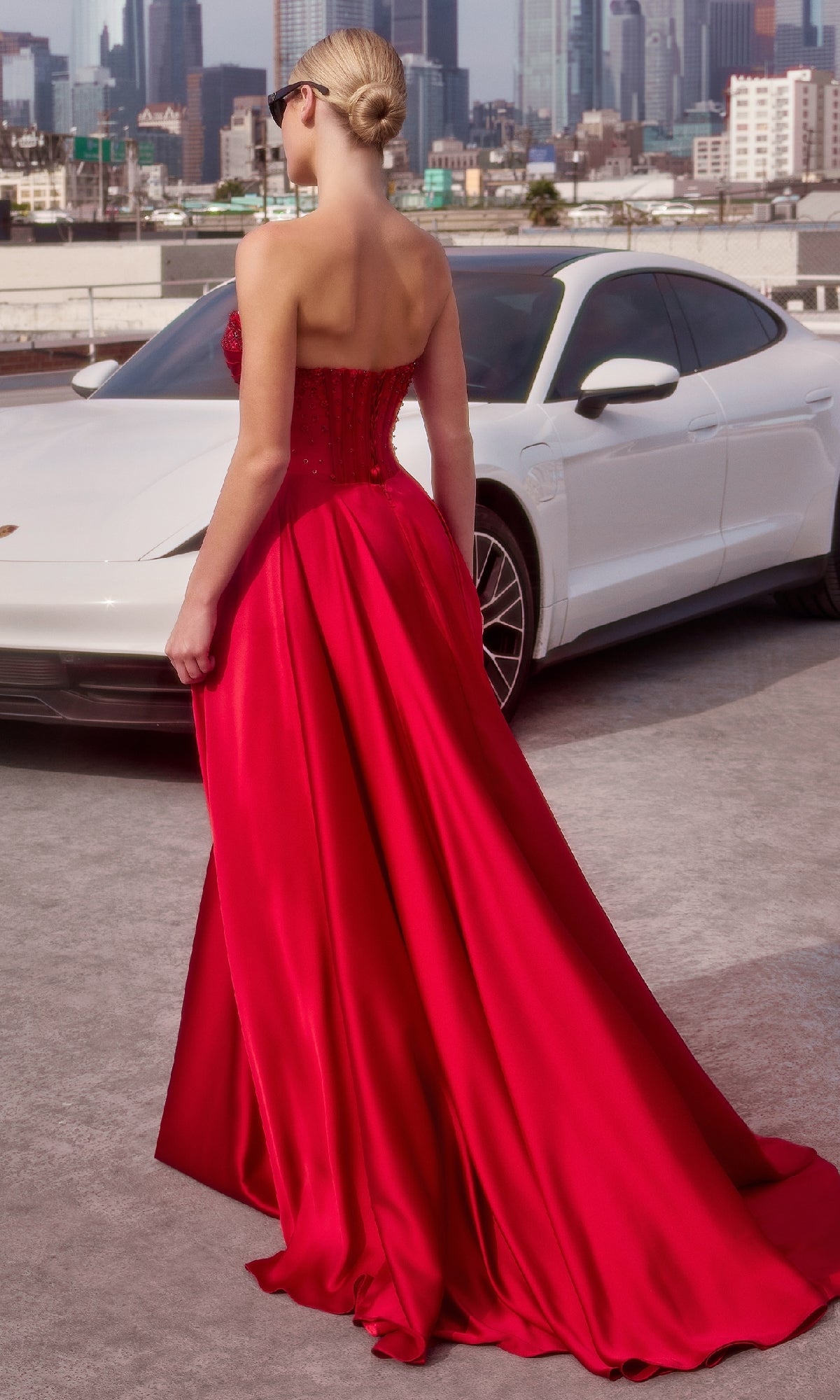 A woman in the Ladivine CD297 Strapless Long A-Line Prom Dress stands on a rooftop near a white car, her elegant gown flowing with the city skyline in the background.