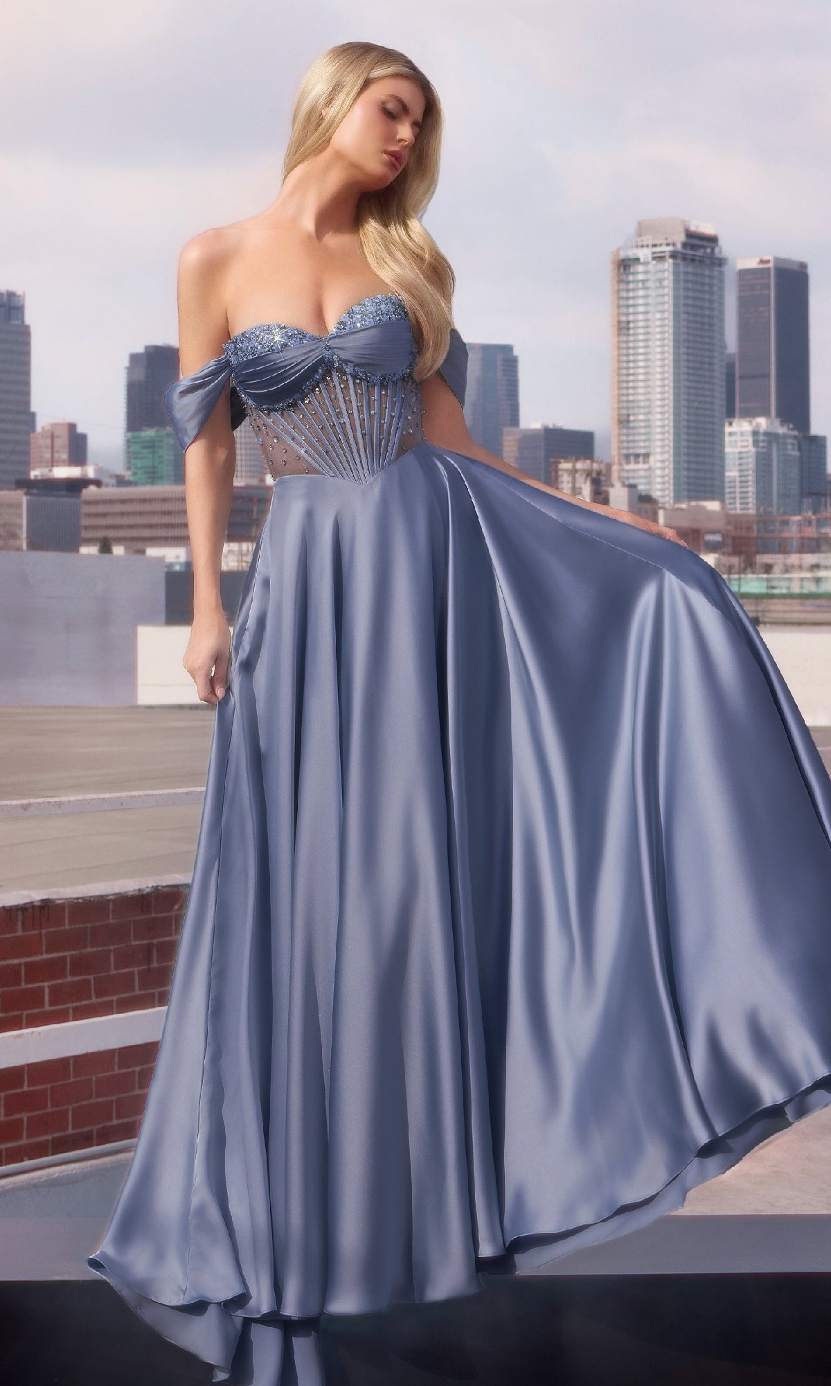 A woman in the Ladivine CD297 strapless long A-line prom dress, featuring a light blue satin skirt and beaded corset, stands on a rooftop with the city skyline glimmering behind her.