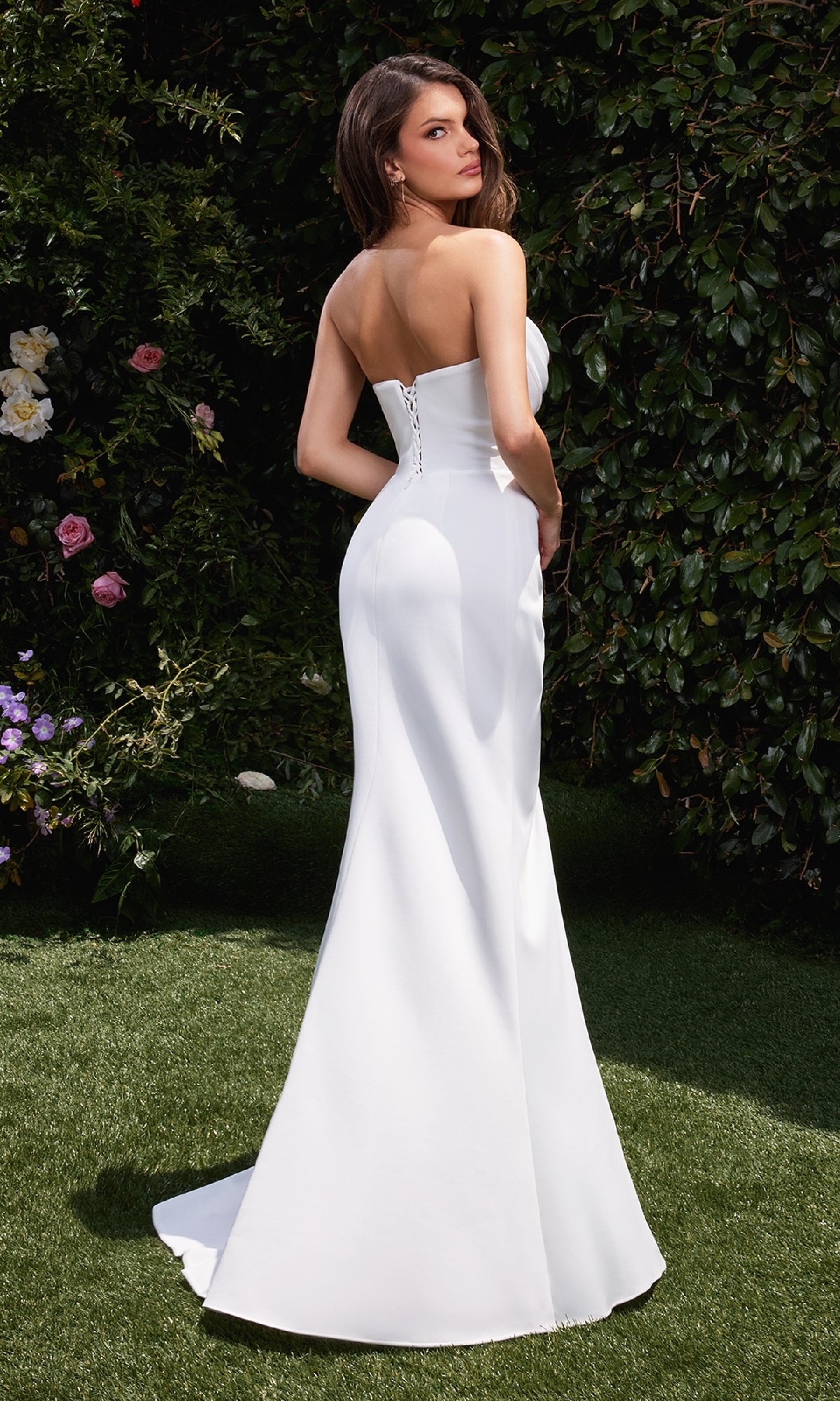 A woman in the Strapless Off-White Prom Gown: Ladivine CD355W stands on grass outdoors, facing slightly away, with greenery and flowers in the background.