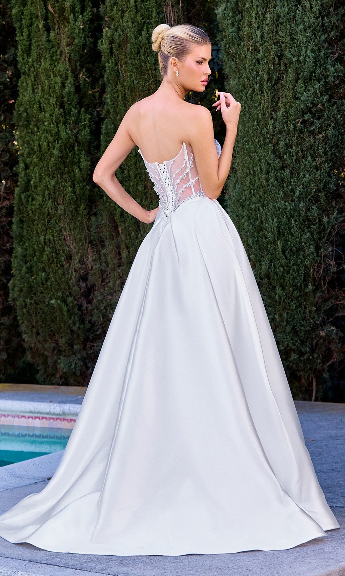 A woman wearing the Ladivine CD389W long white prom dress with a strapless sheer corset bodice and Mikado skirt stands outdoors by a pool and tall green hedges.