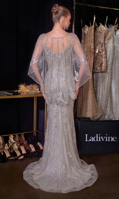 A woman wearing the Ladivine CD637 Long Prom Dress, featuring silver beading and a sheer cape, stands with her back to the camera near a rack of dresses and a shelf of high-heeled shoes.