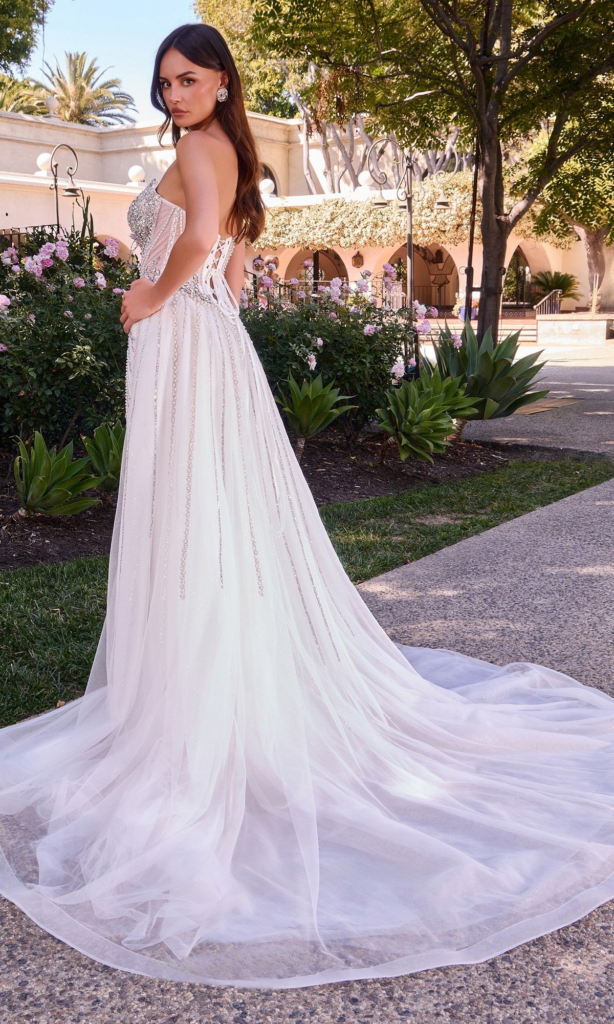 Wearing the Ladivine CD643W, a long formal dress featuring an off-white hue, strapless beaded bodice, and a removable overskirt, a woman stands on a garden path surrounded by blooming flowers and greenery as her elegant tulle train trails behind.
