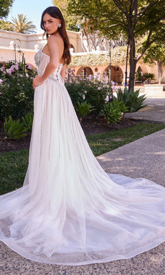 Wearing the Ladivine CD643W, a long formal dress featuring an off-white hue, strapless beaded bodice, and a removable overskirt, a woman stands on a garden path surrounded by blooming flowers and greenery as her elegant tulle train trails behind.