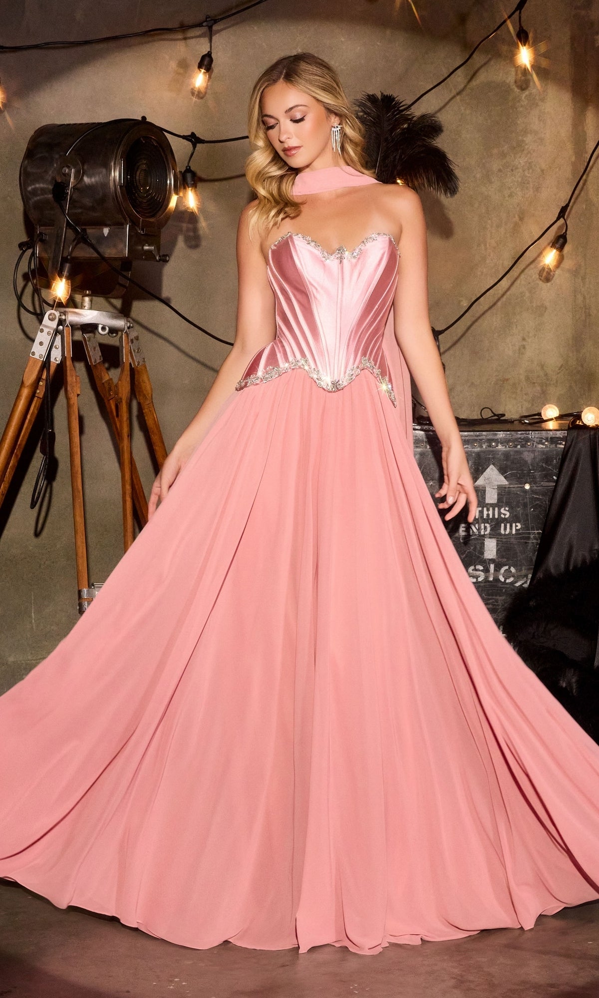 A woman wearing the Ladivine CD668 long prom dress in Dusty Rose, featuring a strapless satin corset bodice and a flowing chiffon A-line skirt, stands indoors against a backdrop of string lights and vintage props.