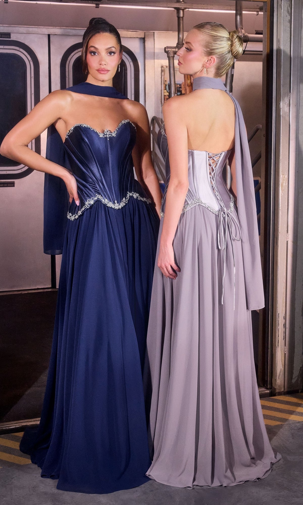 Two women stand side by side in a subway car, both dressed elegantly; one wears the Ladivine CD668 Long Prom Dress in navy, which features a flowing chiffon A-line skirt, while the other dons a strapless light gray gown with a lace-up back.