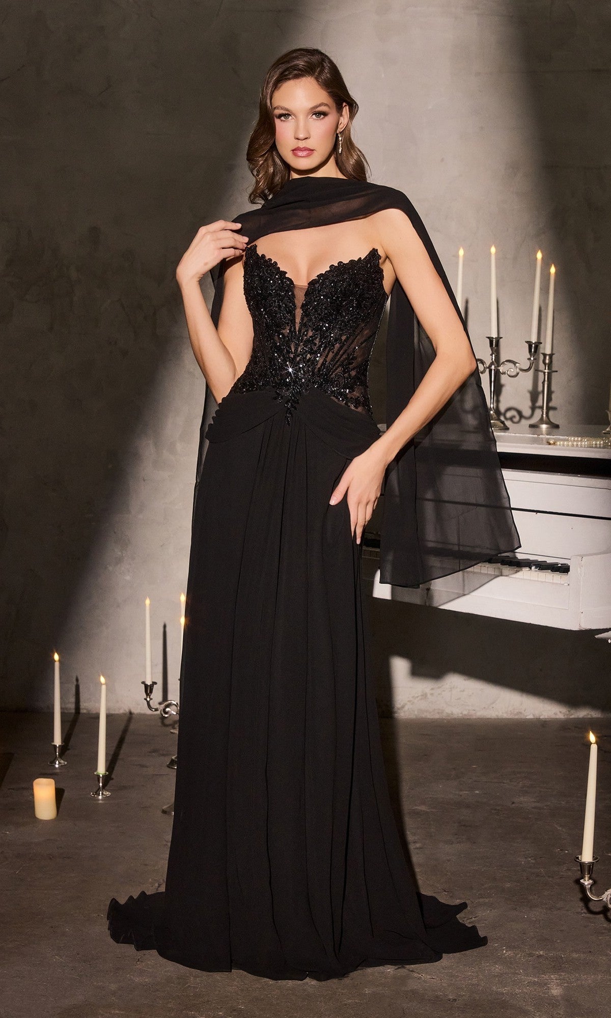 A woman stands gracefully by a white piano with glowing candles, wearing the Ladivine CD673 long black prom dress featuring a strapless beaded corset bodice and sheer details, draped in a matching shawl.