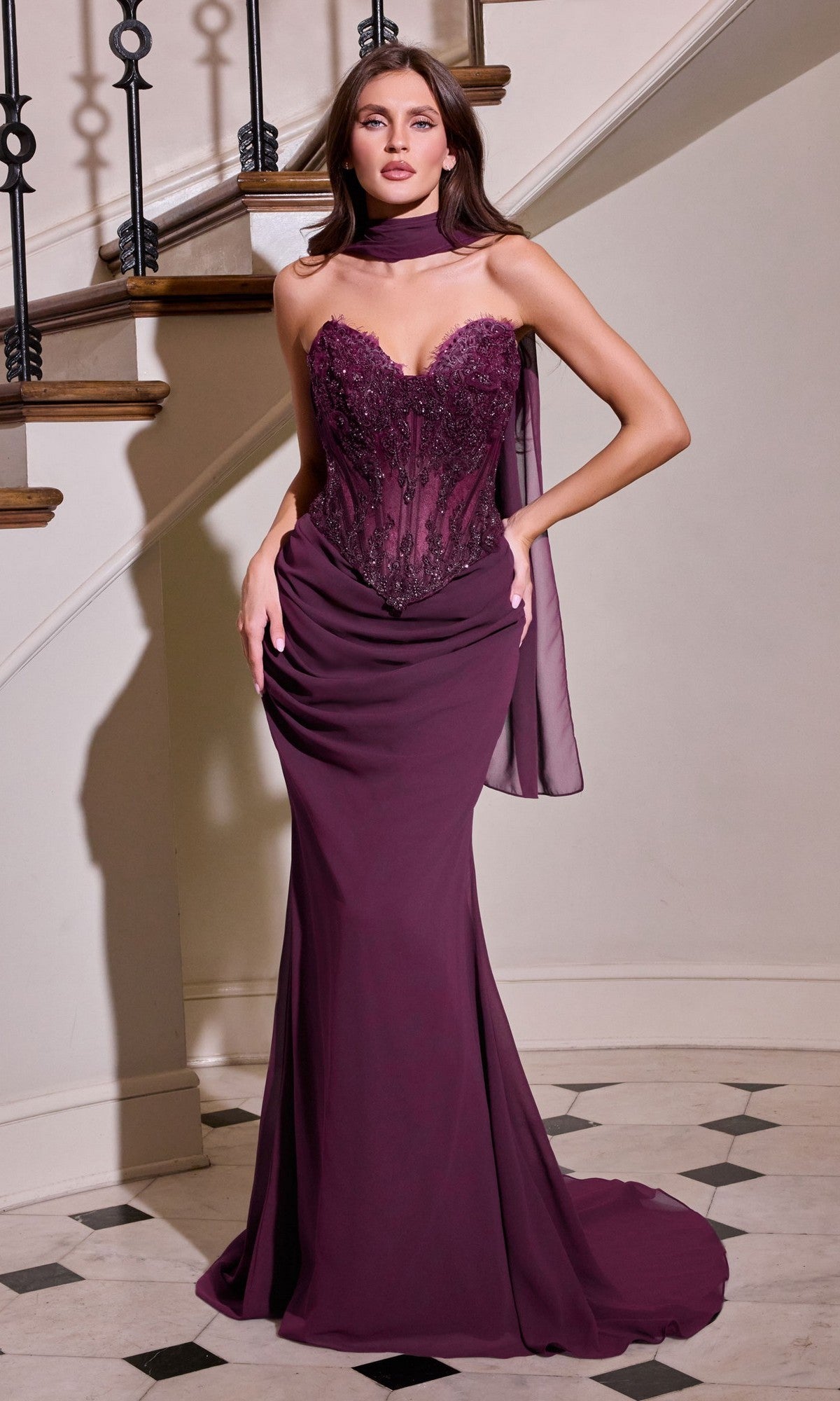 A woman poses gracefully indoors before a curved staircase, wearing the Ladivine CD683 long prom dress in eggplant—a strapless, beaded corset evening gown with a sheer bodice and matching scarf.