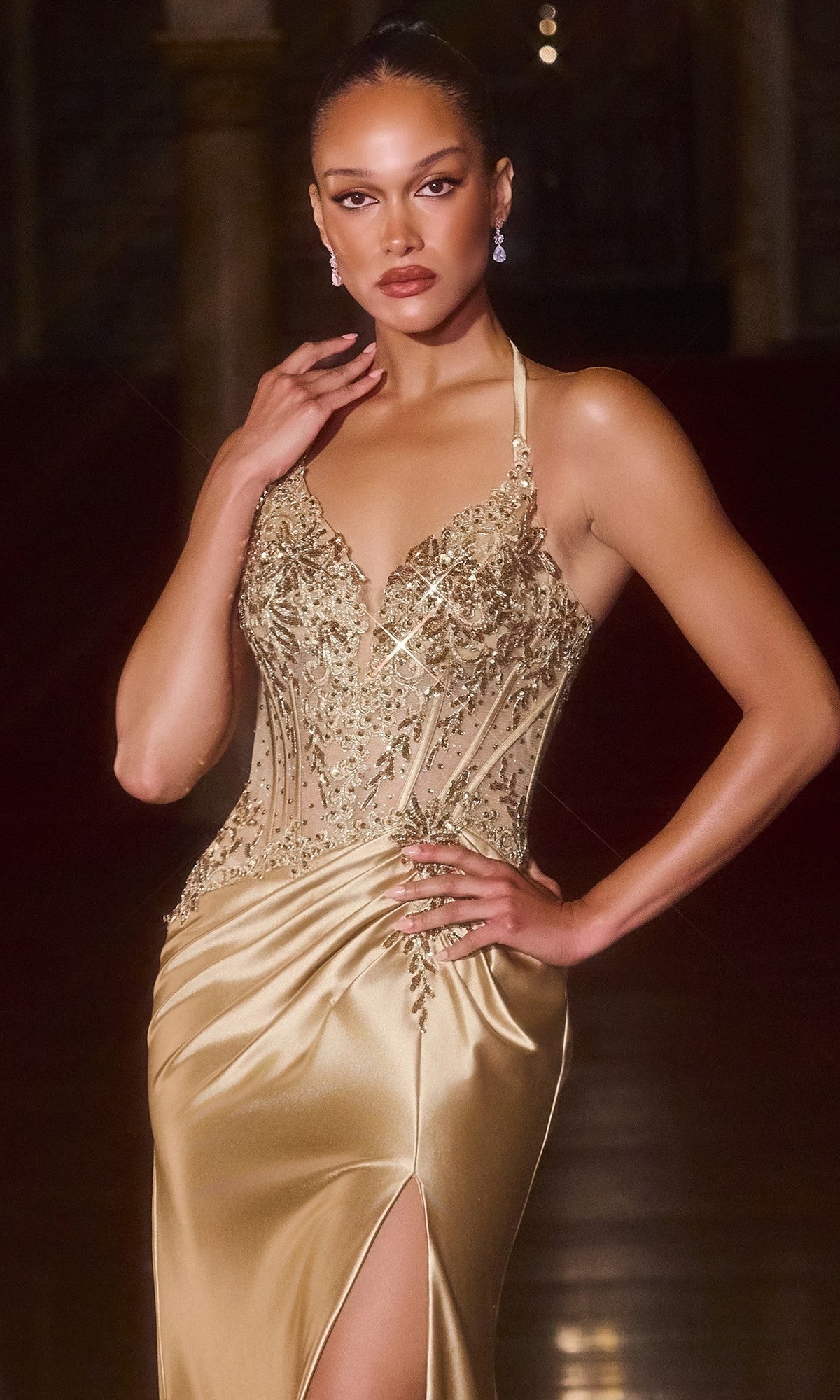 A woman in the Sheer-Corset Long Halter Prom Gown (Ladivine CD690) with an embellished corset-style halter bodice and thigh-high slit poses elegantly indoors, one hand on her hip and the other near her face.