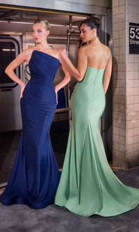 Two women in elegant long gowns—one wearing the Ladivine CD694 asymmetric strapless prom gown in navy—stand on a subway platform by a 53rd Street sign, capturing city glamour.