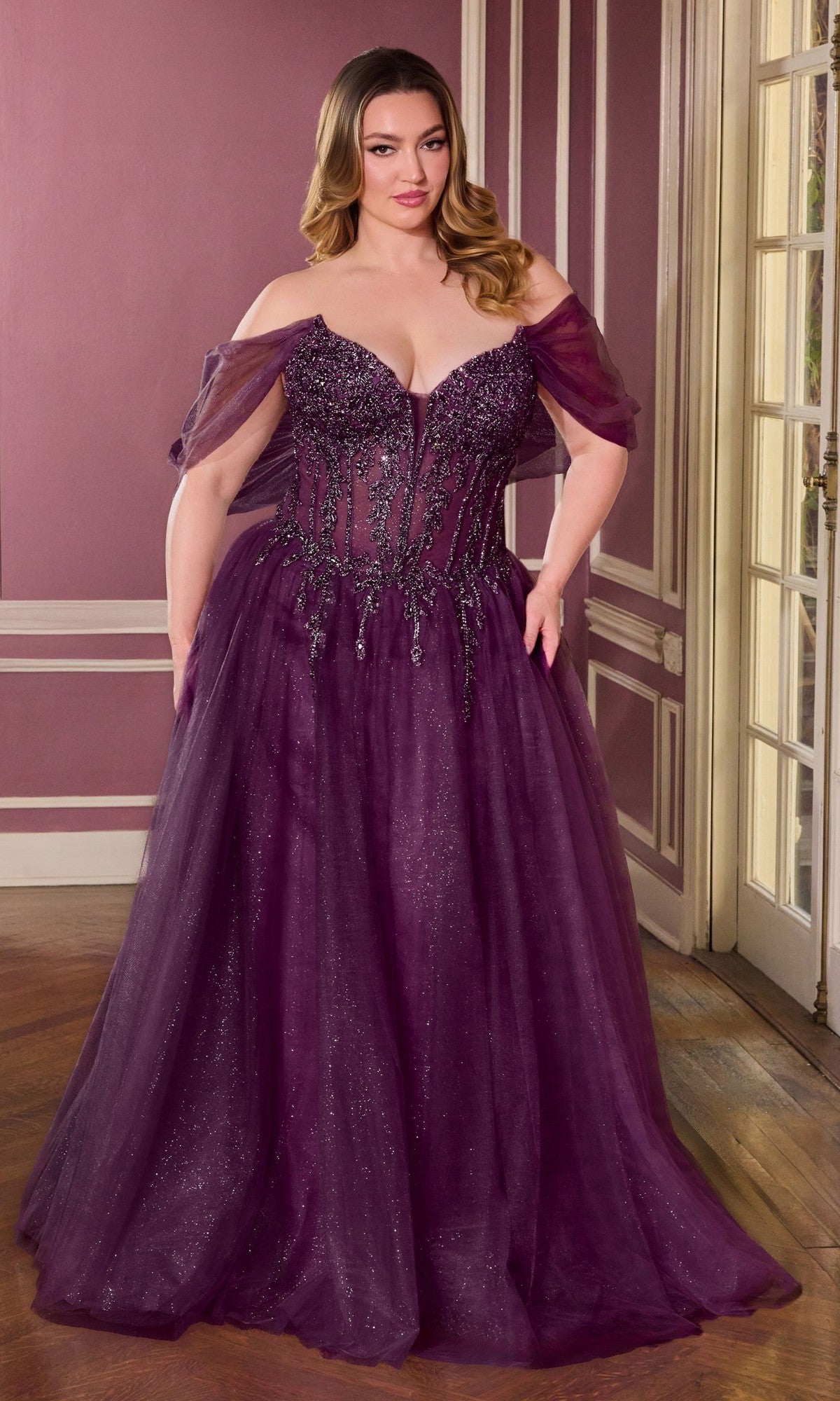 A woman poses gracefully indoors in the Ladivine CD721C long plus-size prom dress, featuring an embellished corset bodice and off-the-shoulder sleeves in elegant eggplant, standing on a wooden floor before paneled walls and a glass door.