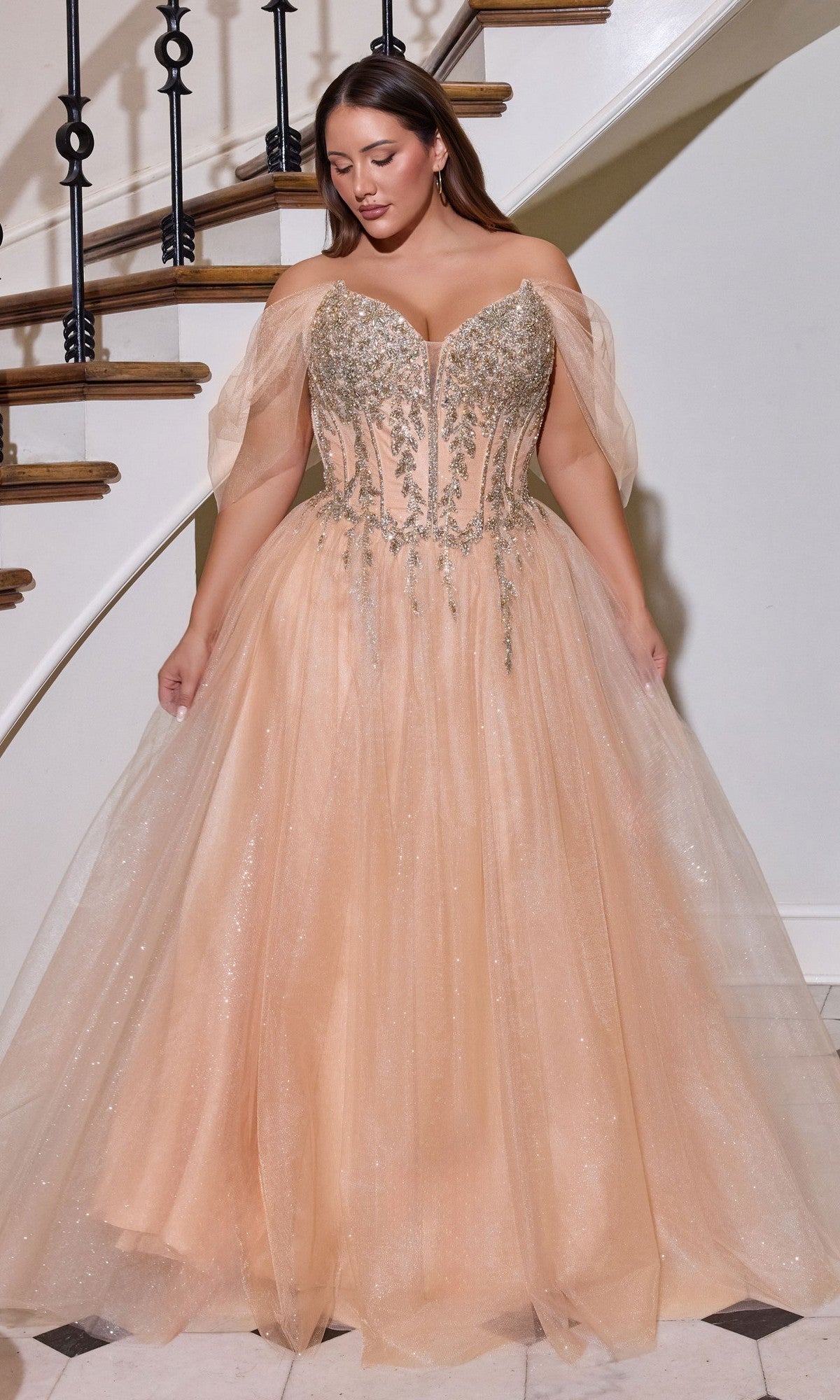 A woman stands indoors on tiled flooring, wearing the gold Ladivine CD721C long plus-size prom dress with an embellished corset bodice and glitter tulle ball gown skirt, in front of a curved staircase.